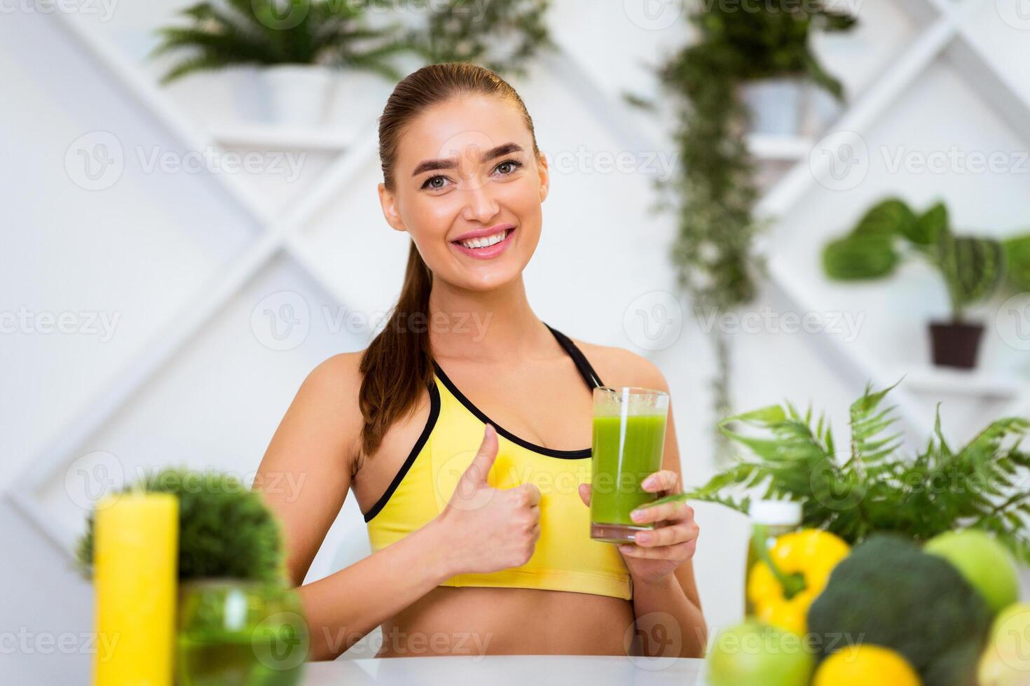 A woman in a yellow sports bra holds a glass of green smoothie while giving a thumbs up. Shes surrounded by green plants and fruits, in a kitchen setting. photo