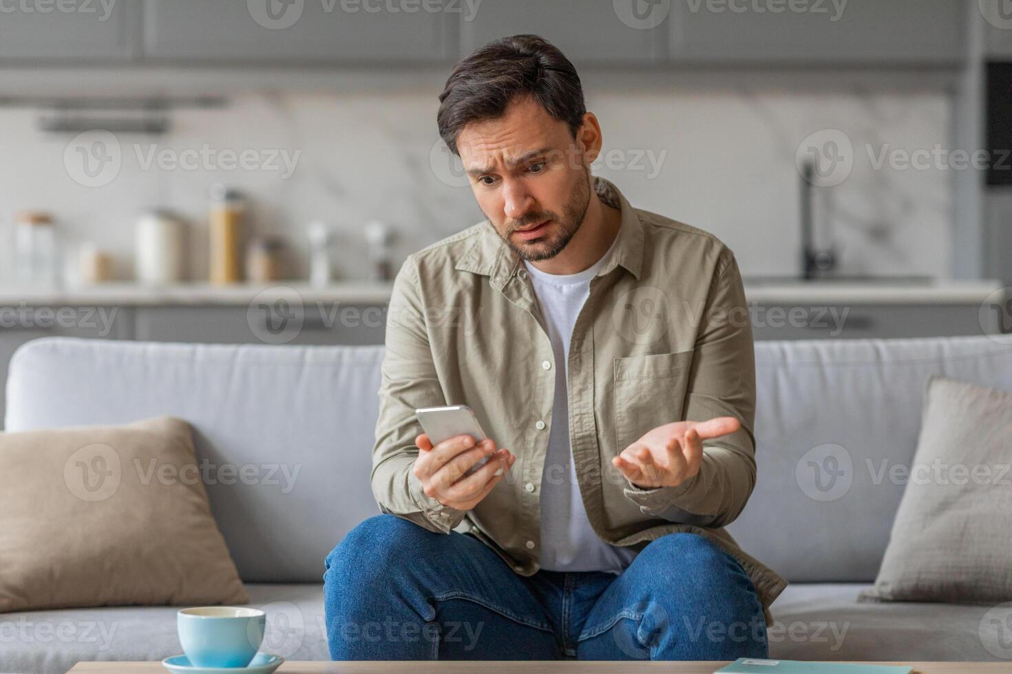 A man sits on a couch in his living room, holding his phone with a confused expression on his face. He gestures with his other hand, as if trying to explain or clarify something. photo