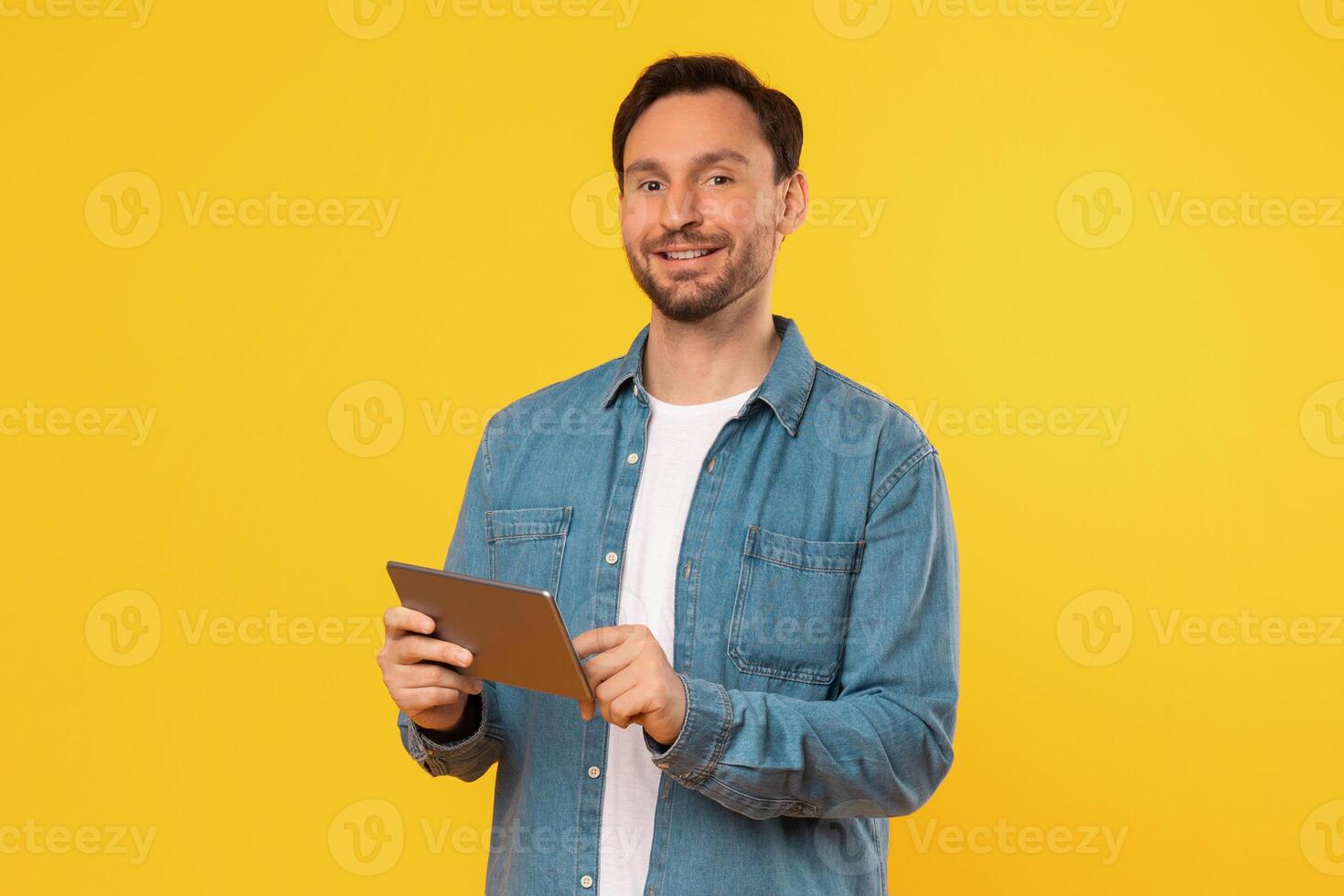 A man in a blue denim shirt stands in front of a yellow background. He is smiling and holding a tablet in his hands. photo