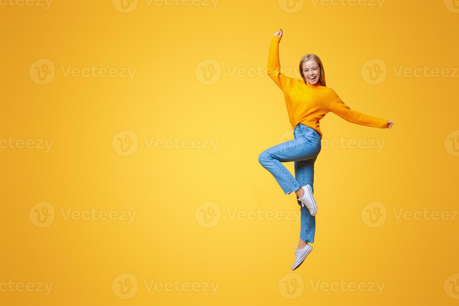 Hooray. Happy young girl jumping in studio and looking at camera on orange background with copy space photo