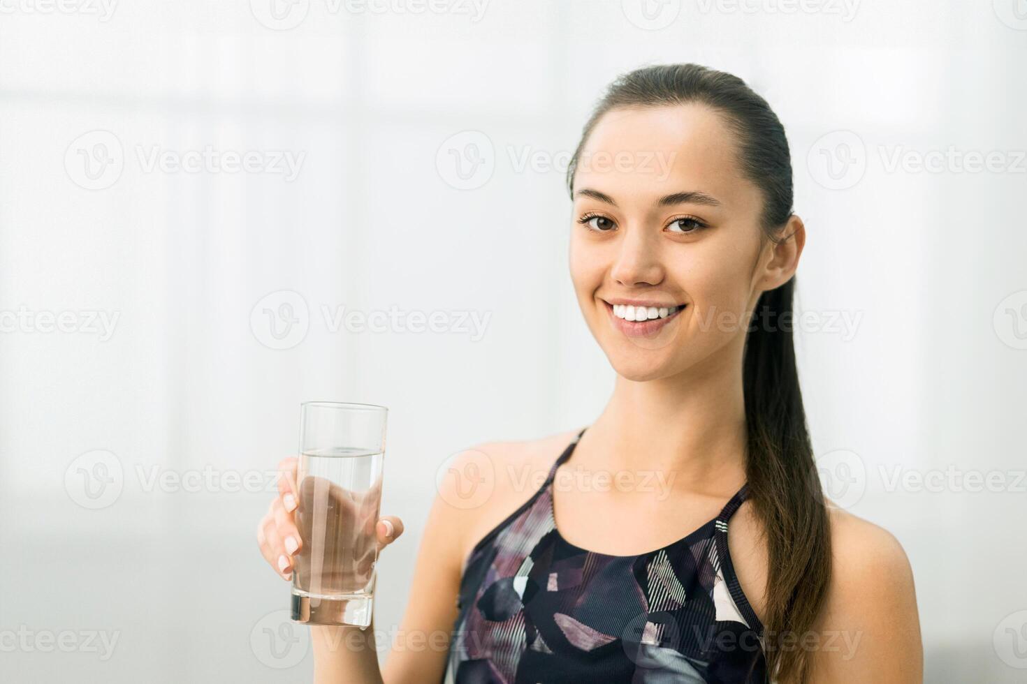A woman in athletic wear holds a glass of water, smiling happily after a workout. photo