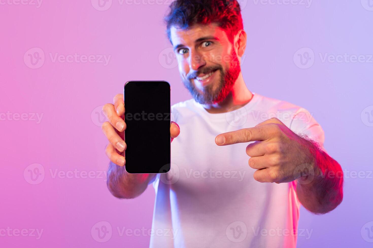 A cheerful man with a beard, wearing a white t-shirt, is excitedly pointing at a smartphone blank screen while standing against a vibrant pink and purple neon background. photo