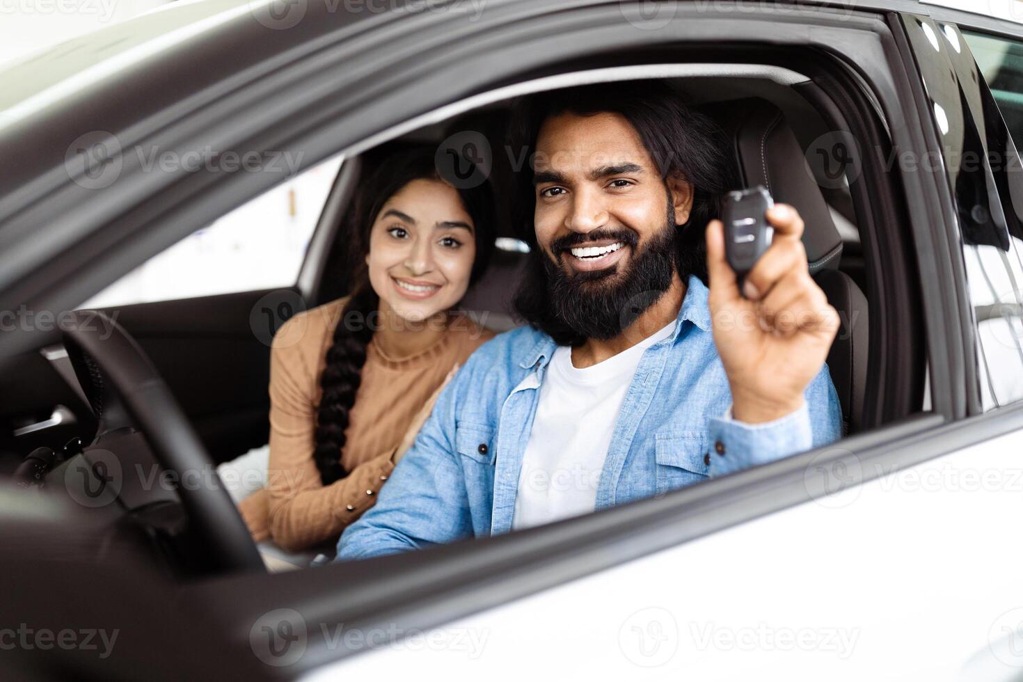 A cheerful and excited Indian couple is seated inside a new car. The man, with a beard, holds up car keys, indicating they have likely just purchased the vehicle, while the woman smiles warmly photo