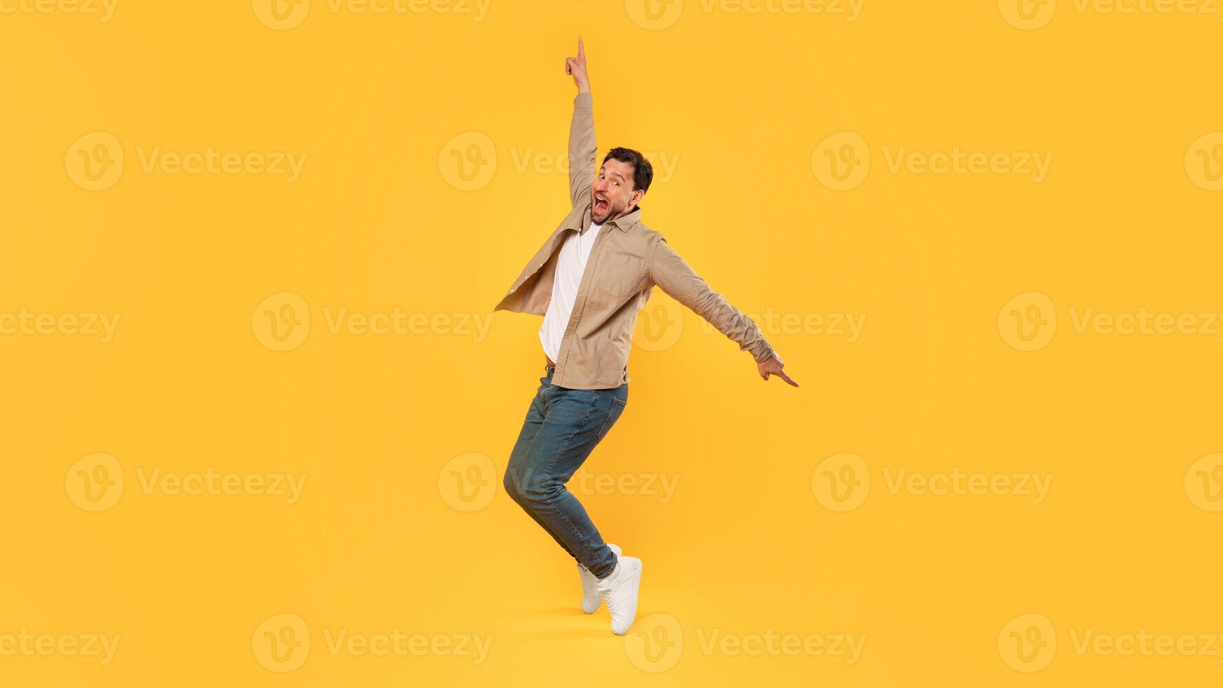 A man with a beard and a tan jacket jumps in the air with his right arm raised, pointing upward. He is wearing blue jeans and white sneakers and is facing the camera. He has a shocked expression photo