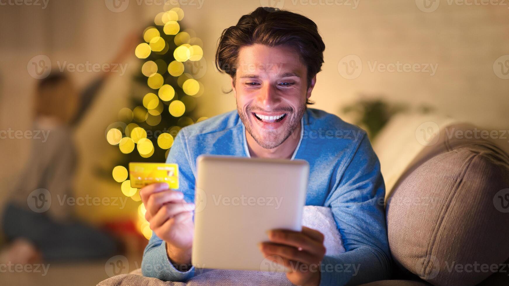 Handsome man shopping online on tablet on New Year's eve, woman decorating Christmas tree on background photo