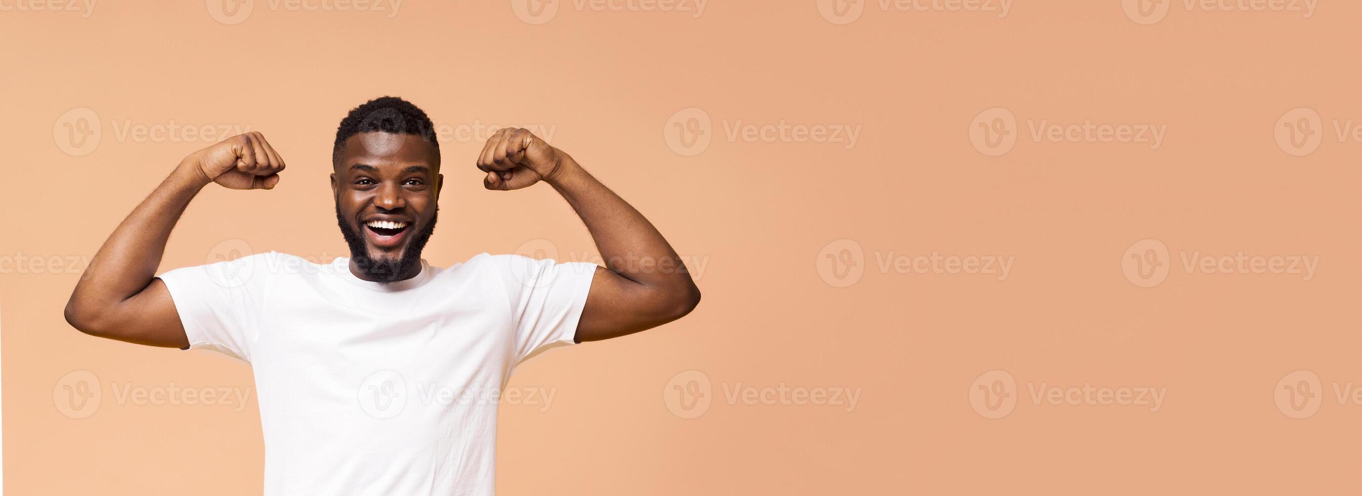 A happy, young, Black man with a short beard is flexing his muscles while standing in front of a beige background. He is wearing a white t-shirt and has a big smile on his face, copy space, panorama photo