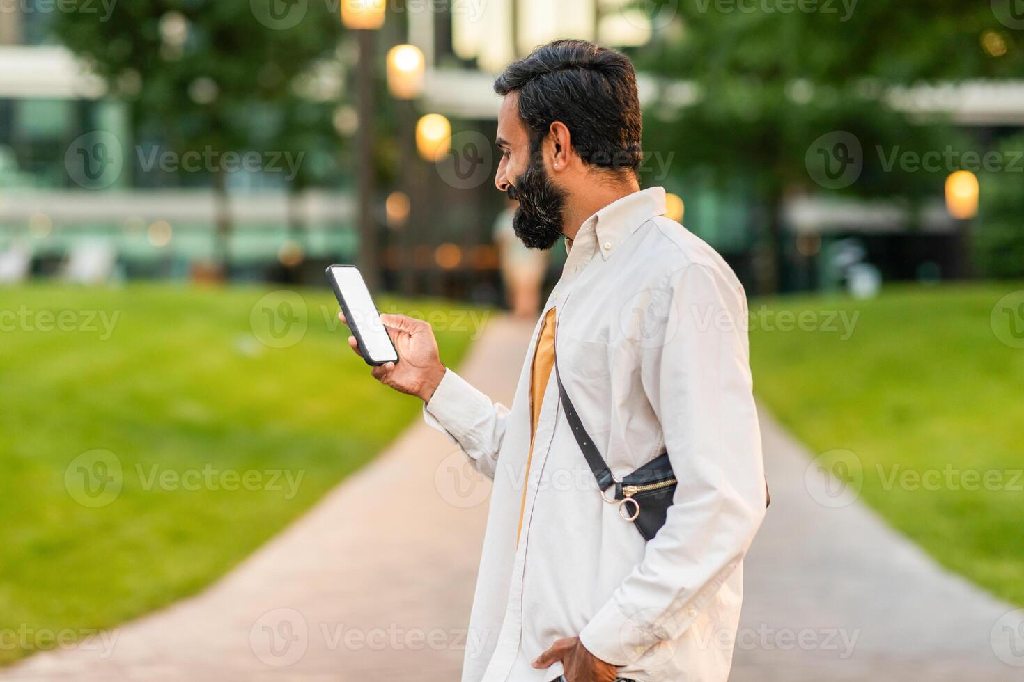 Indian man dressed in a light-colored shirt walks down a pathway in a vibrant urban park during the early evening. He is attentively looking at his smartphone, possibly checking messages or browsing. photo