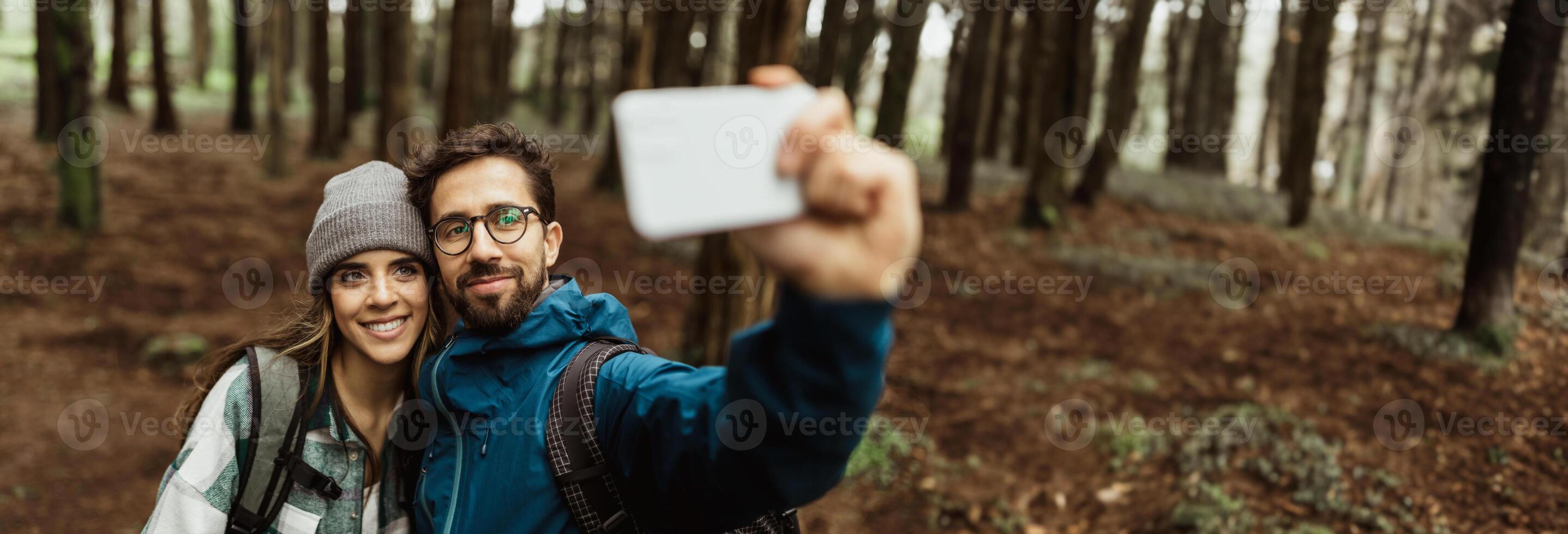 A couple is taking a selfie in a forest. The man is holding the phone and the woman is smiling at the camera. They are both wearing casual clothing and backpacks, copy space photo