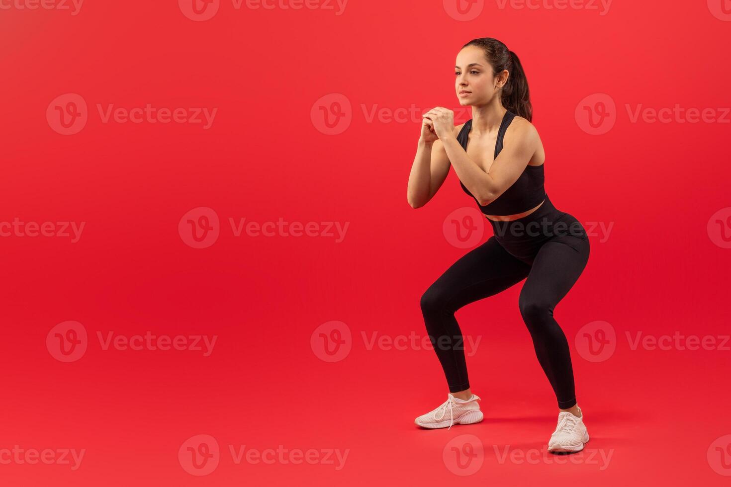 A focused young woman wearing a black sports outfit with white sneakers is engaged in a squatting exercise. Her hands are clenched together in front of her as she maintains a proper form, copy space photo