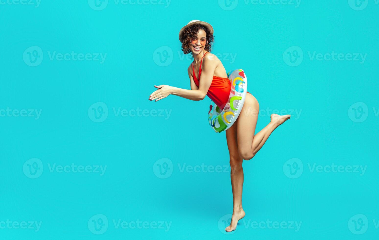 A happy black woman with curly hair is wearing a red swimsuit and a straw hat. She is holding an inflatable ring in one hand, and she is posing against a blue background. photo