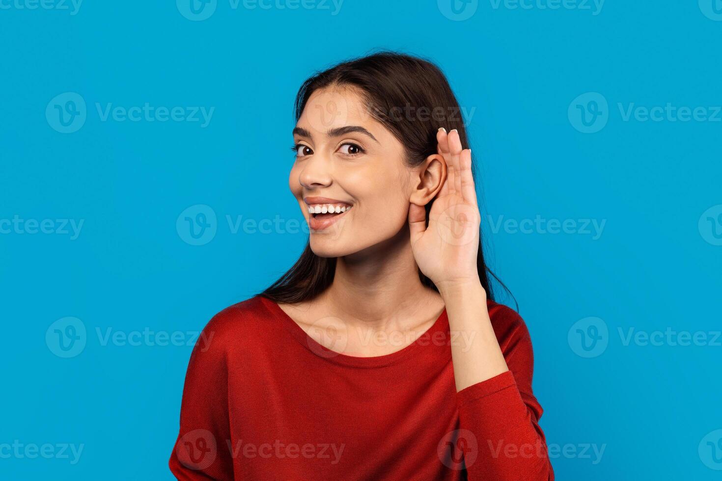 A woman with a joyful expression on her face is smiling and touching her ear with her hand, indicating she might be listening or adjusting an earpiece. She appears content and engaged in the moment. photo