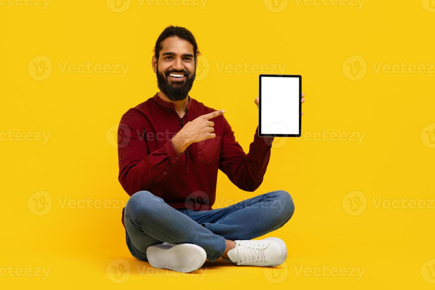 Indian man is seated on the floor, holding a tablet with white blank screen in his hands, isolated on yellow studio background photo