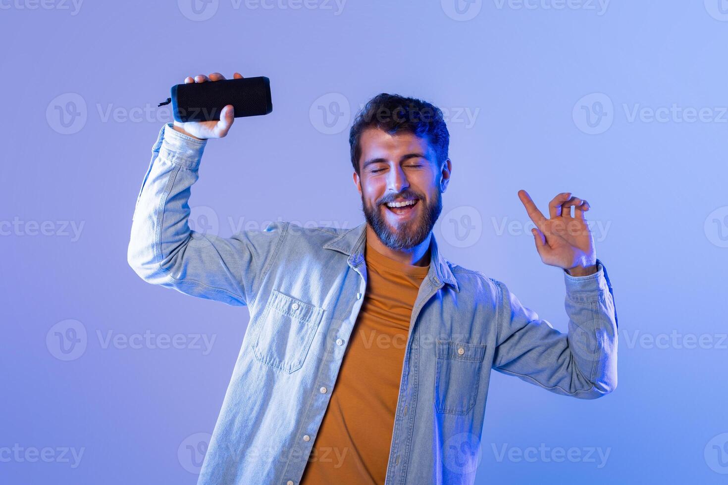 A young bearded man in a casual denim jacket is dancing joyfully with Bluetooth speaker in his hand. The background is illuminated with blue and purple lighting photo
