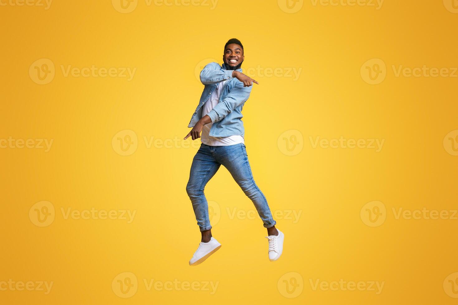 Look there. Funny african american man jumping in air and pointing at copy space over yellow studio background. photo