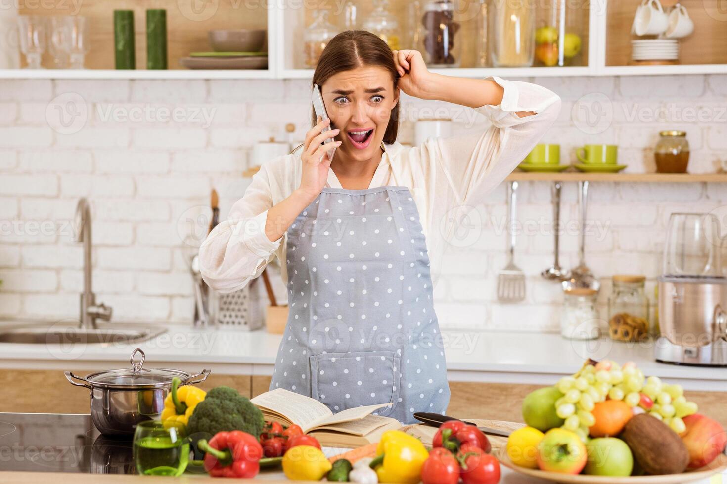 A woman in a kitchen, wearing an apron, speaks on the phone with a shocked expression. She appears to have received bad news. The kitchen counter is filled with fresh vegetables and fruits. photo