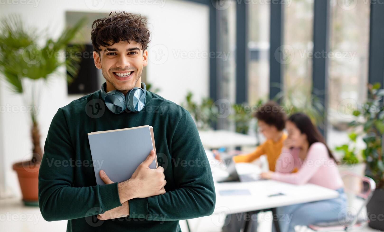 European man college student holding books and smiling at camera while female classmates studying with laptop on the background, coworking space photo