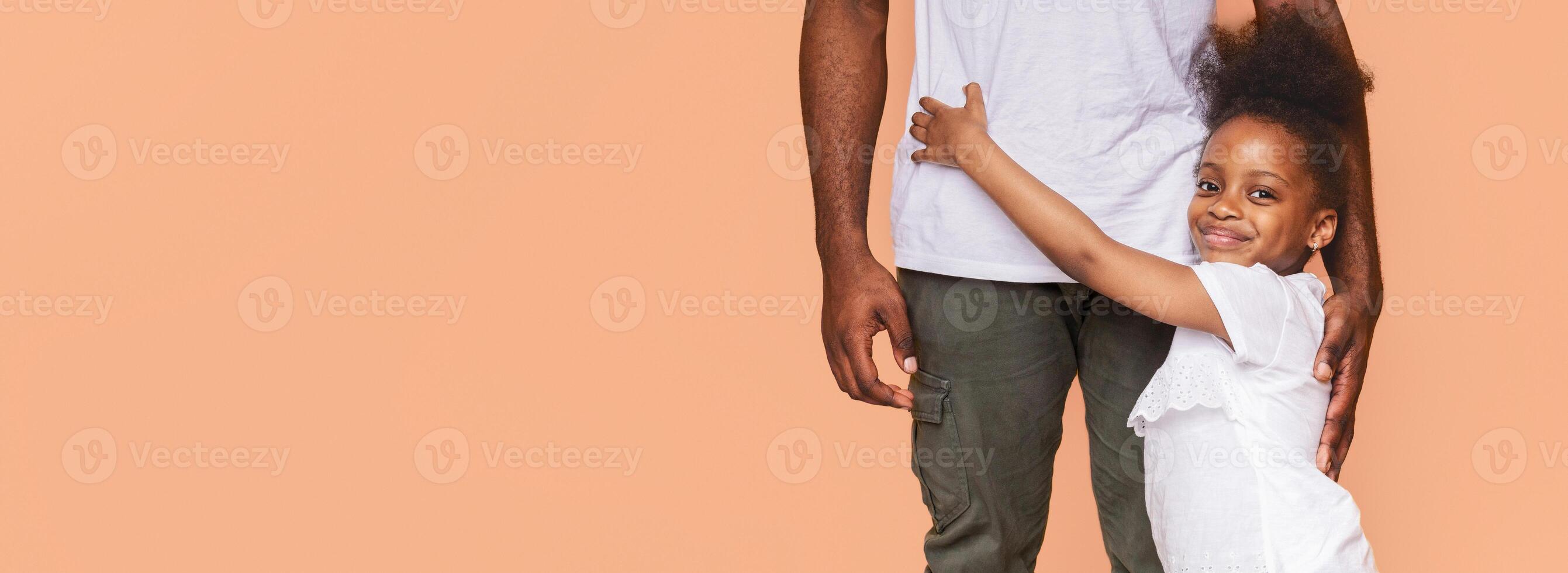 A little black girl with dark curly hair hugs her fathers waist from behind, looking at the camera and smiling with joy. Both are wearing white shirts against a light peach background. photo