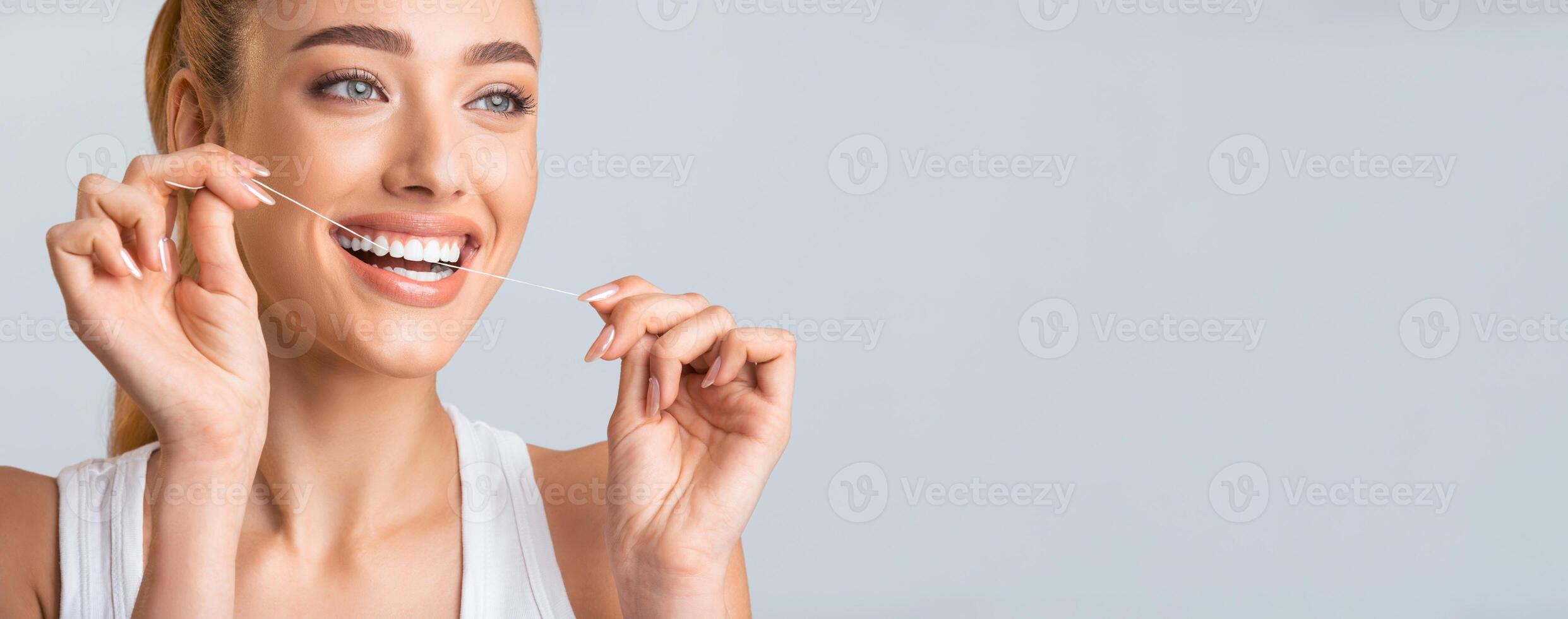 A young woman with blonde hair smiles brightly as she flosses her teeth. Her white teeth are clearly visible, and she is looking towards the left side of the frame, panorama with copy space photo