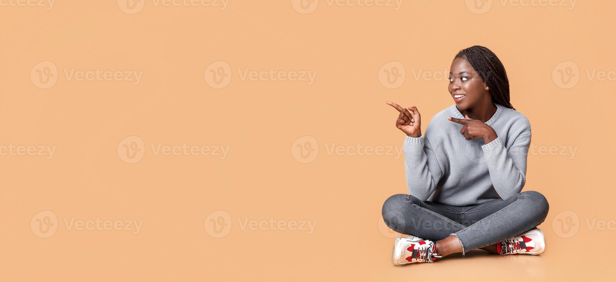 Happy african woman in blue sitting on floor and pointing at copy space, beige background, panorama photo