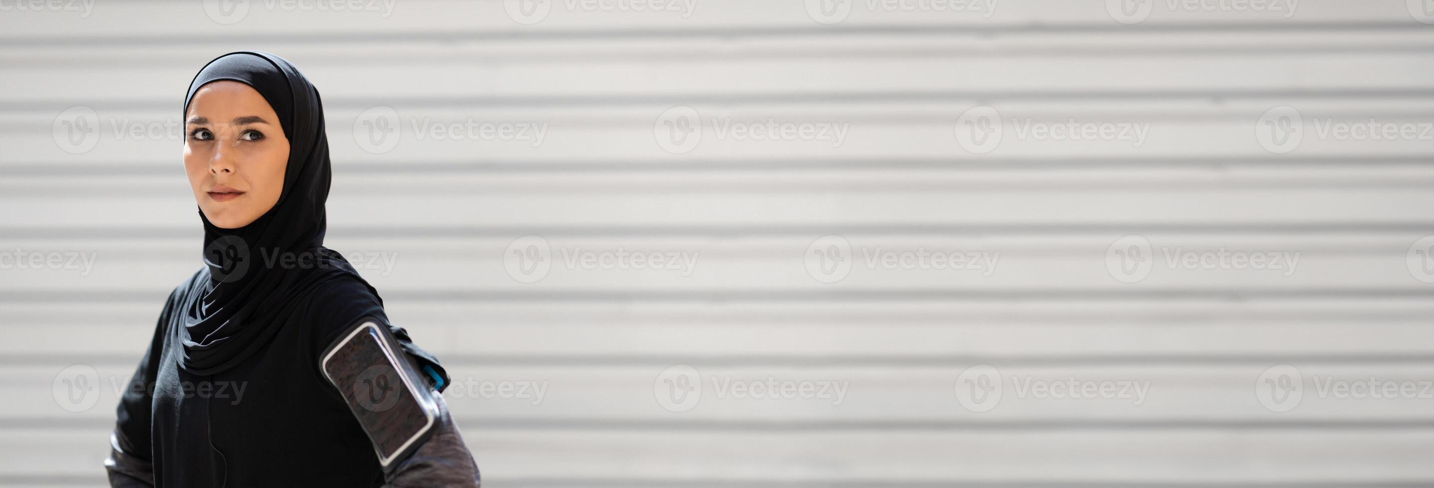 Confident serious calm pretty young muslim woman in hijab with phone on her shoulder resting from exercises on gray wall background. Outdoor sports, body care in city, training, jogging, copy space photo