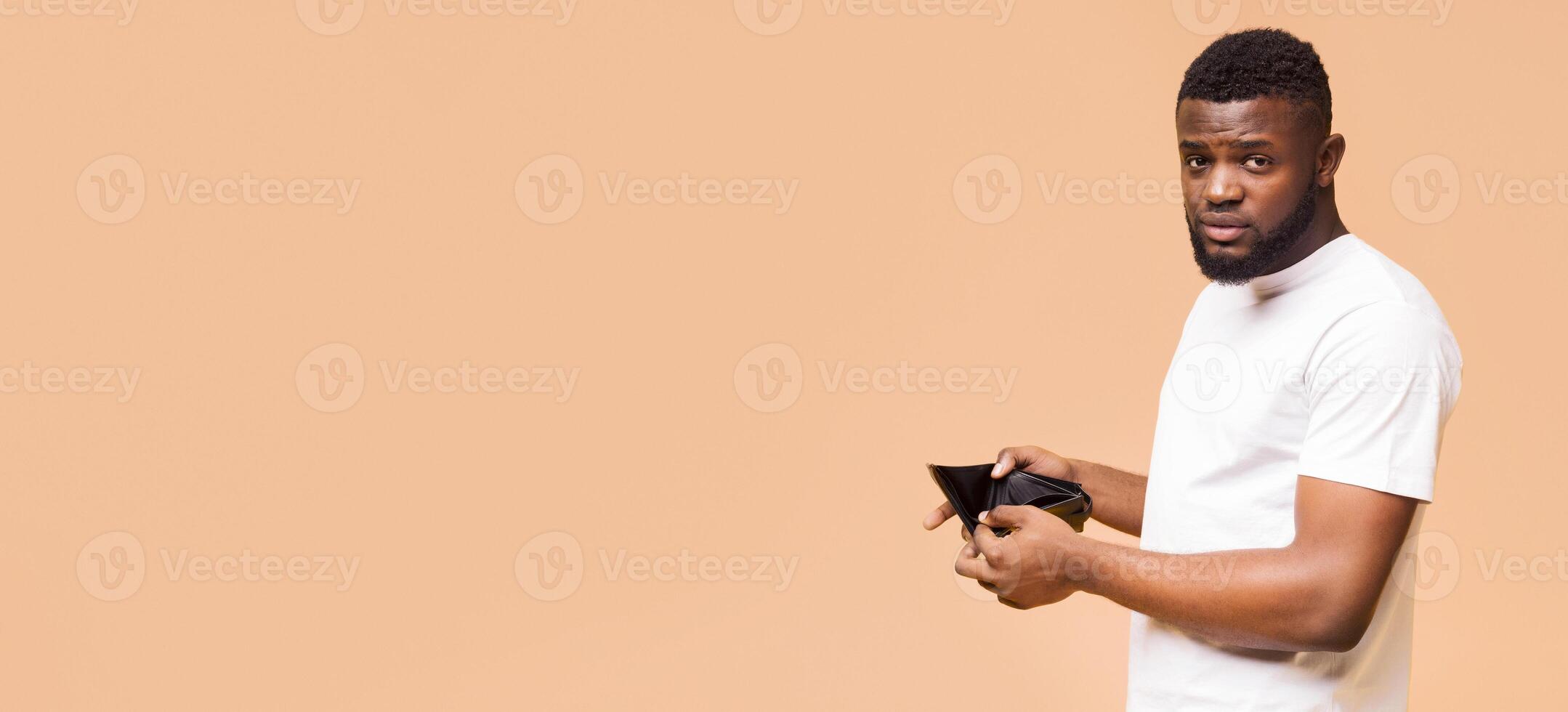 A young African American man in a white shirt stands in front of a beige background, holding his empty wallet open and looking towards the camera with a concerned expression, copy space photo