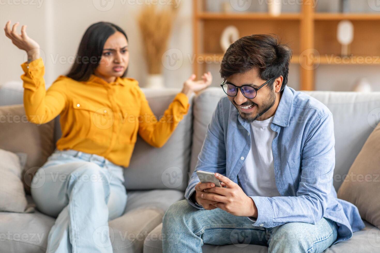 Indian man wearing glasses and a blue shirt is smiling and looking at his phone. Next to him, a woman in a yellow shirt is sitting with raised hands, displaying frustration photo