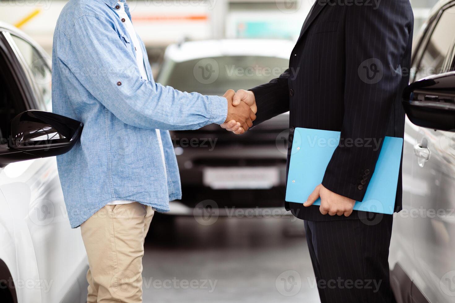 Cropped of two individuals are standing in front of a car, engaged in a handshake gesture. Both men are dressed formally, and the car in the background adds a professional touch to the scene. photo