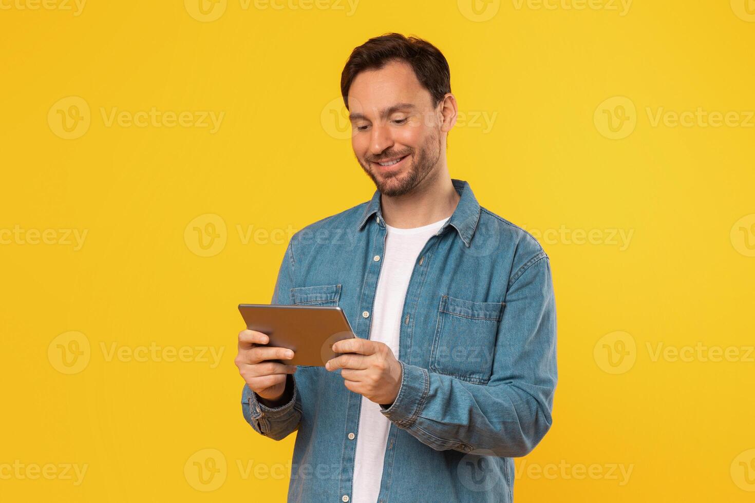 A man wearing a denim shirt stands in front of a bright yellow background, holding a tablet computer in his hands and looking down at the screen. He is smiling as he interacts with the tablet. photo