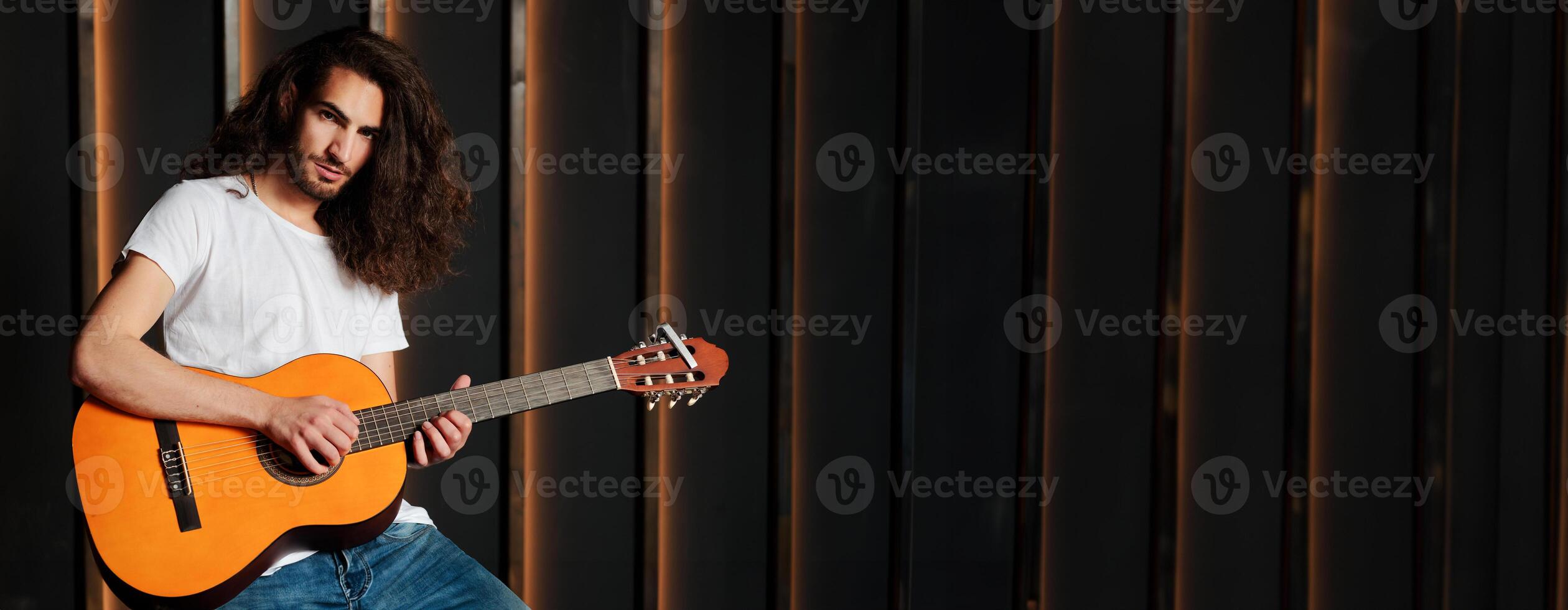 Talented Hispanic Musician Man Playing Acoustic Guitar, Touching Strings Of Musical Instrument And Looking At Camera Posing In Dark Studio Indoor, Standing Near Empty Space For Advertisement photo