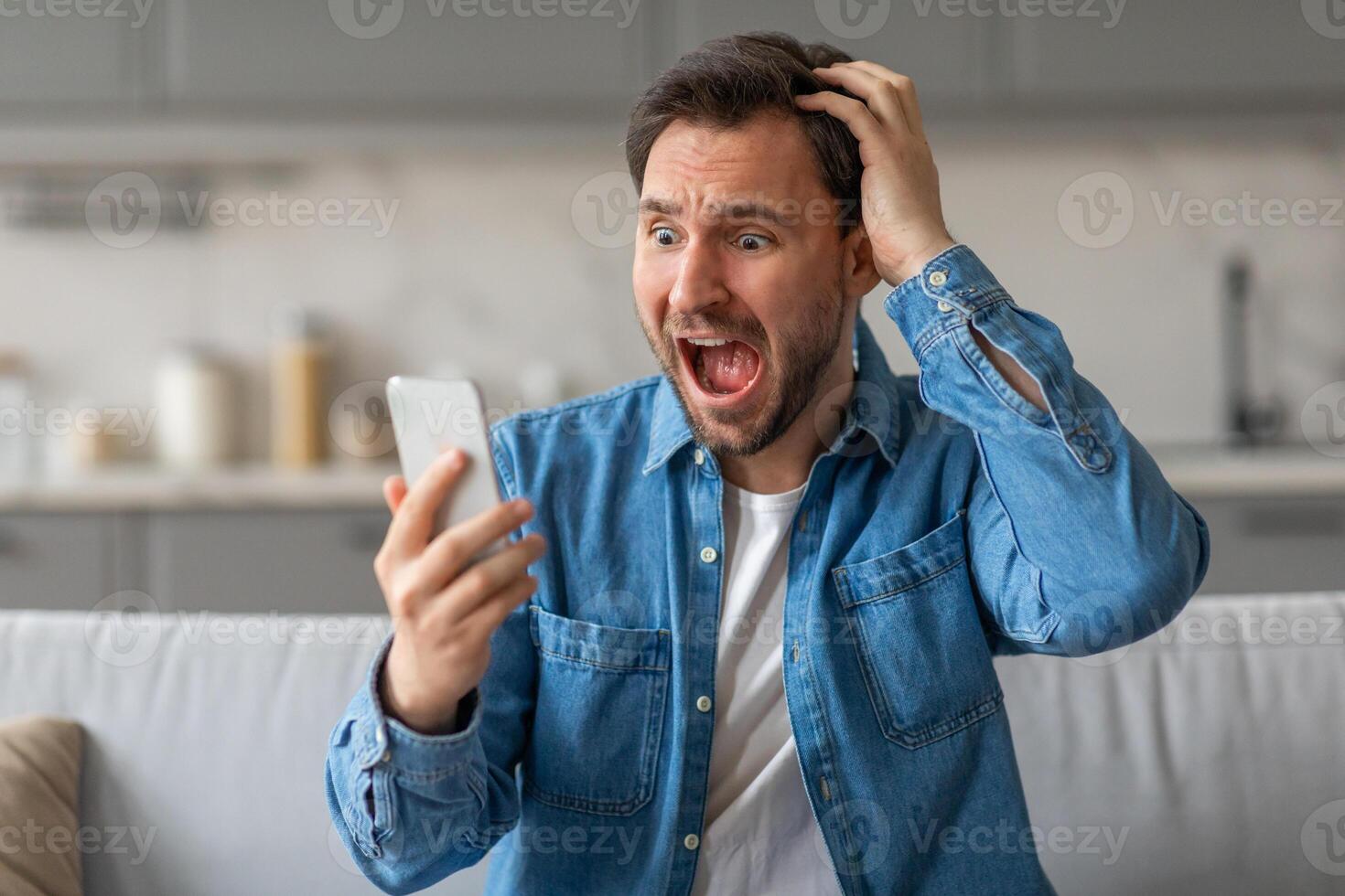 A man in a blue denim shirt is sitting on a couch in a kitchen. His mouth is open in shock, and he has his hand on his head. He is looking at a phone screen in his other hand. photo