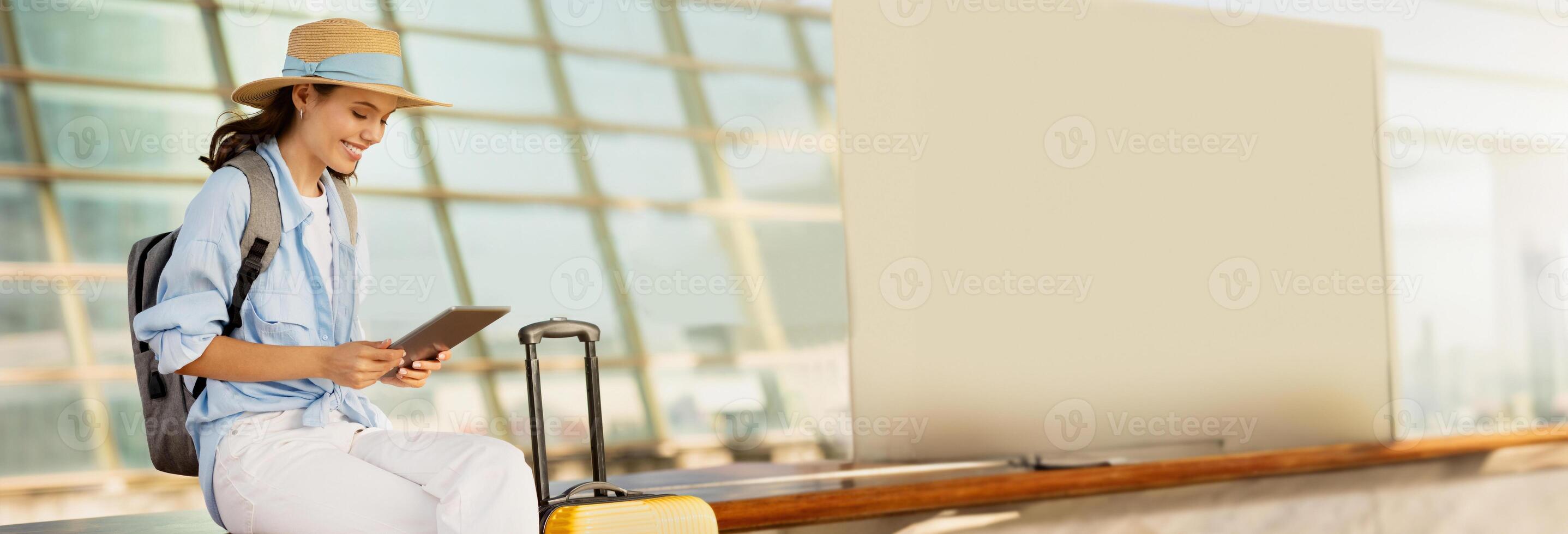Cheerful young european woman in hat with suitcase and backpack, typing on tablet, enjoy journey on station, airport. App for tourist, business trip, active lifestyle, work and vacation, copy space photo