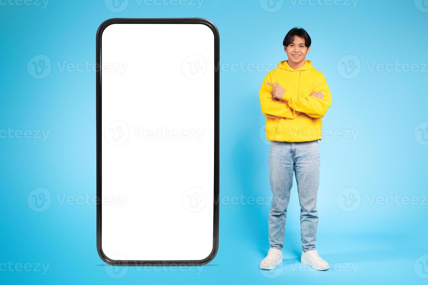 Asian young guy dressed in a yellow hoodie and blue jeans stands with arms crossed next to a large blank smartphone screen. The background is bright blue, creating a clean and modern look. photo