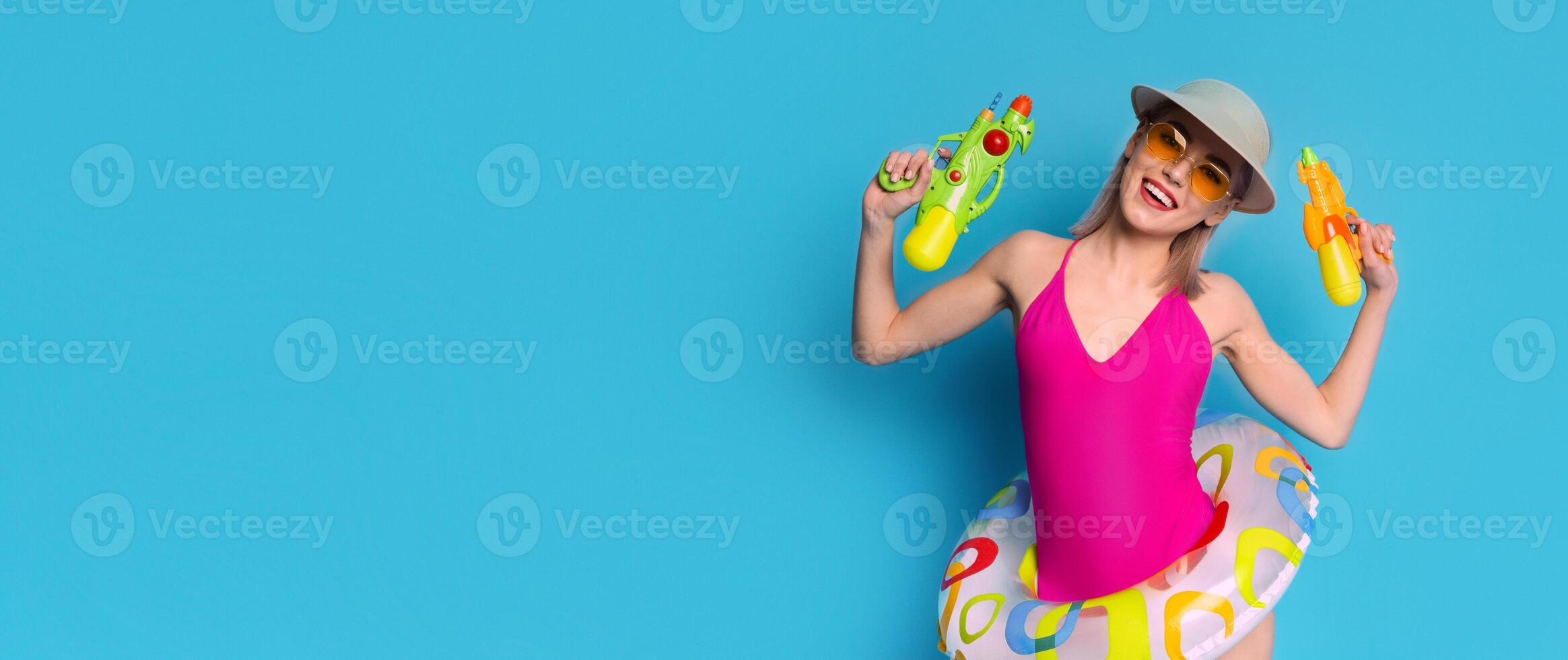 A young woman in a pink swimsuit, a sun hat, and sunglasses holds two water guns. She smiles at the camera with a playful expression. Her joyful demeanor suggests fun and excitement photo
