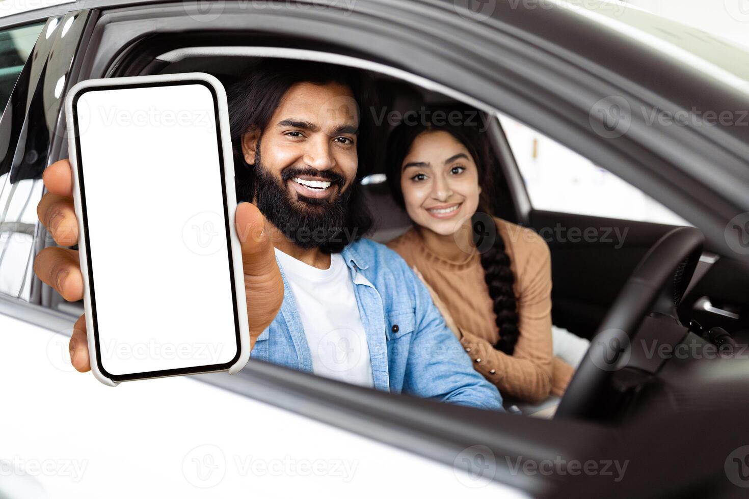 A bearded Indian man is extending his arm out of a car window, holding a smartphone with a blank screen towards the camera possibly for an interactive display or advertising purpose. photo