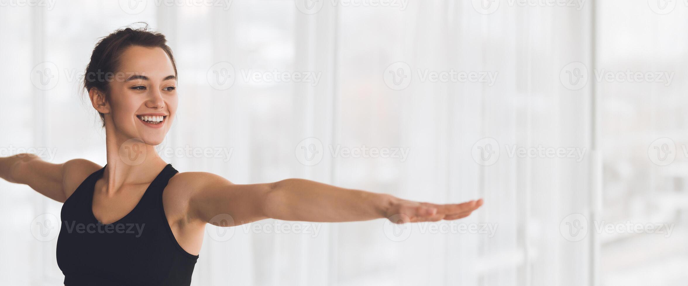 A woman practices yoga in a sunlit studio, extending her arms and smiling as she engages in a pose, showcasing her dedication to wellness, copy space photo