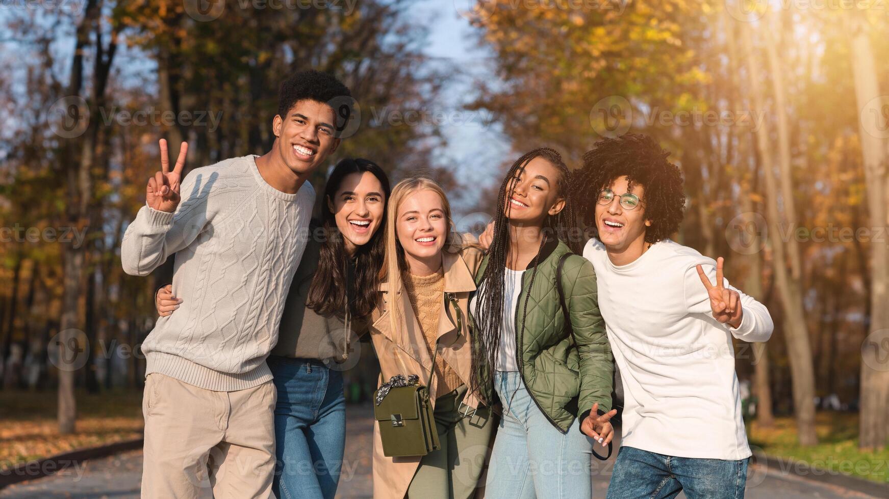 Cheerful multiethnic teenagers guys and girls posing at camera outdoors, showing peace gesture, park background photo