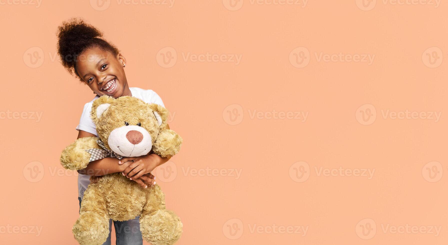 A little Black girl with curly hair smiles at the camera while holding a brown teddy bear against a peach-colored background, copy space photo