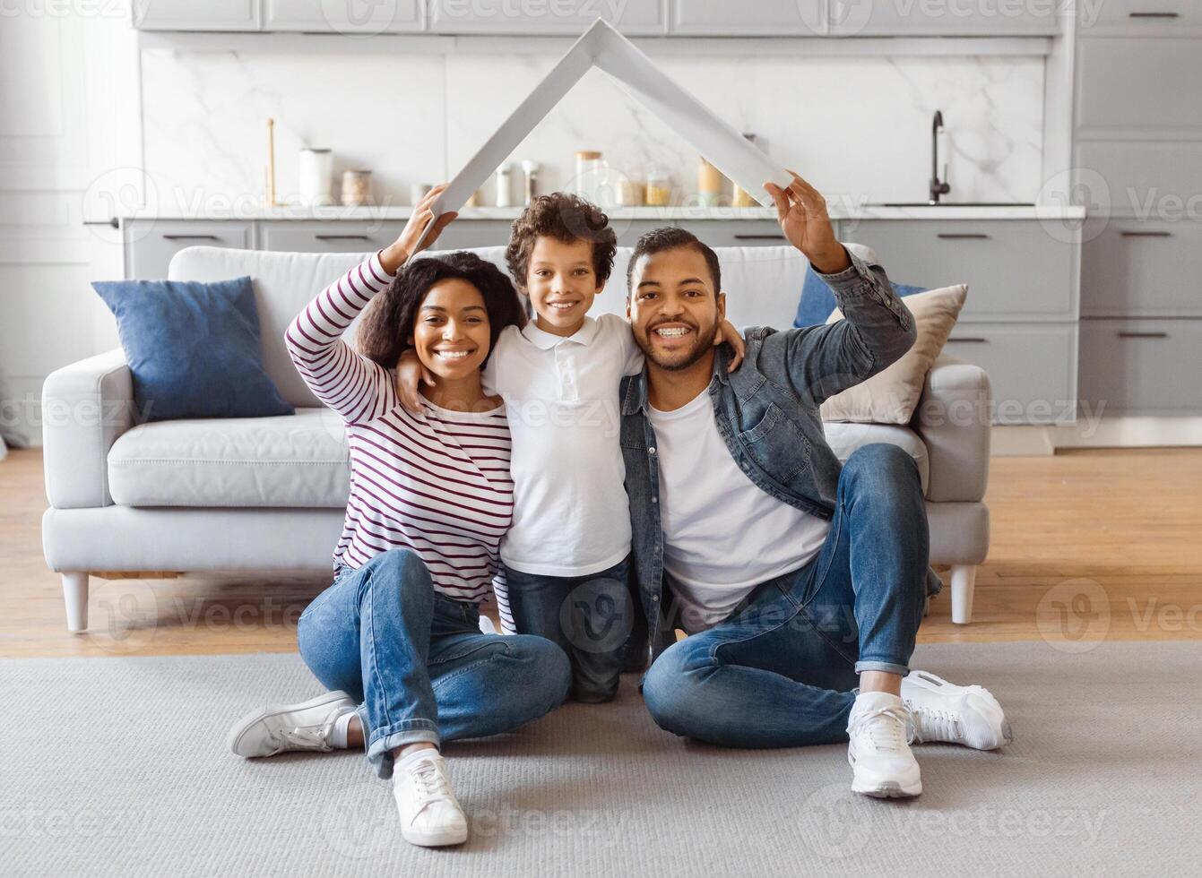 African American family seated on the floor facing a couch. They appear to be engaged in conversation or watching something together. Parents holding carton roof above their child photo