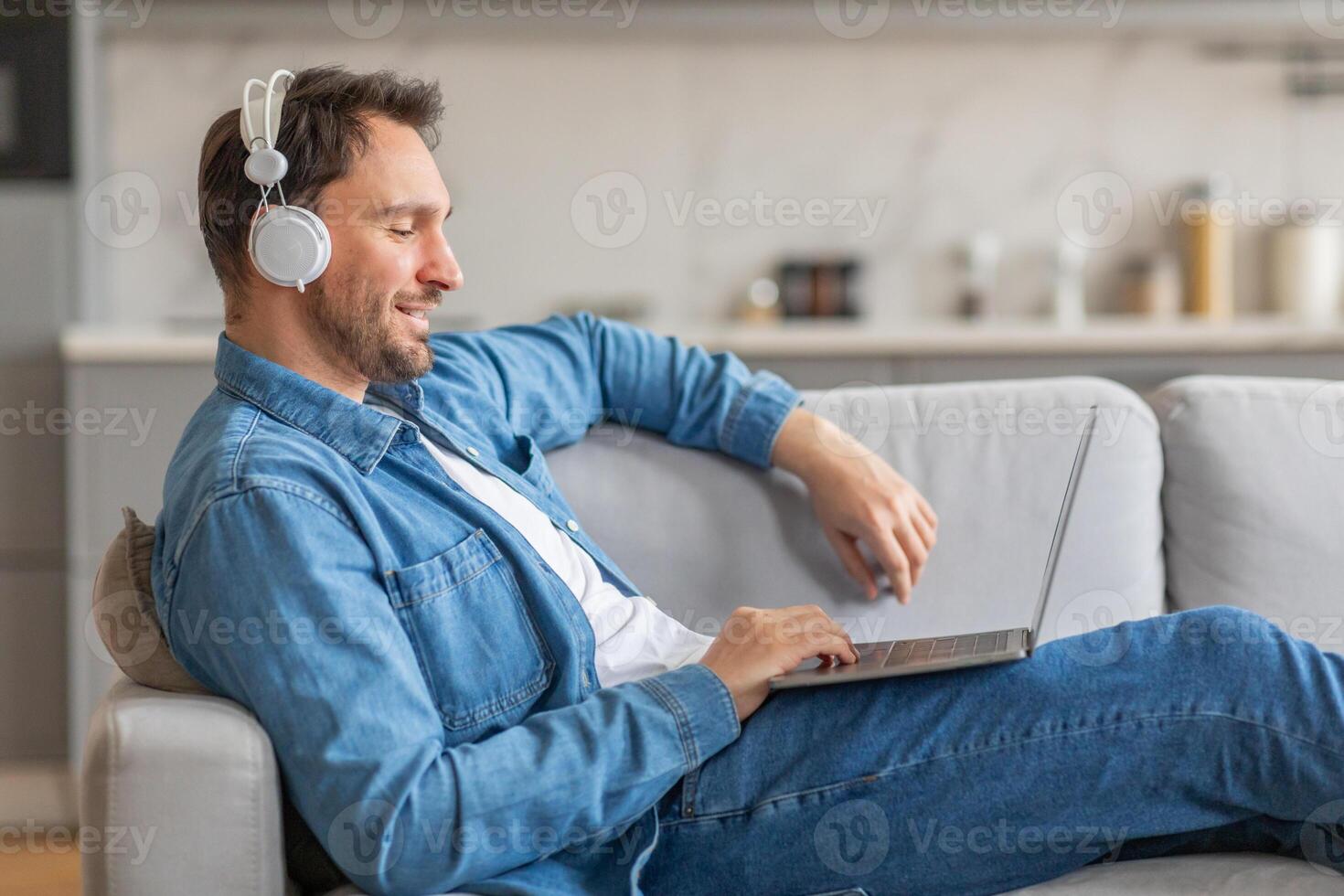 A man in a denim shirt sits on a light-colored couch, relaxed with headphones on and his laptop in his lap. He is smiling, looking at the screen of his computer. The room has a modern design photo