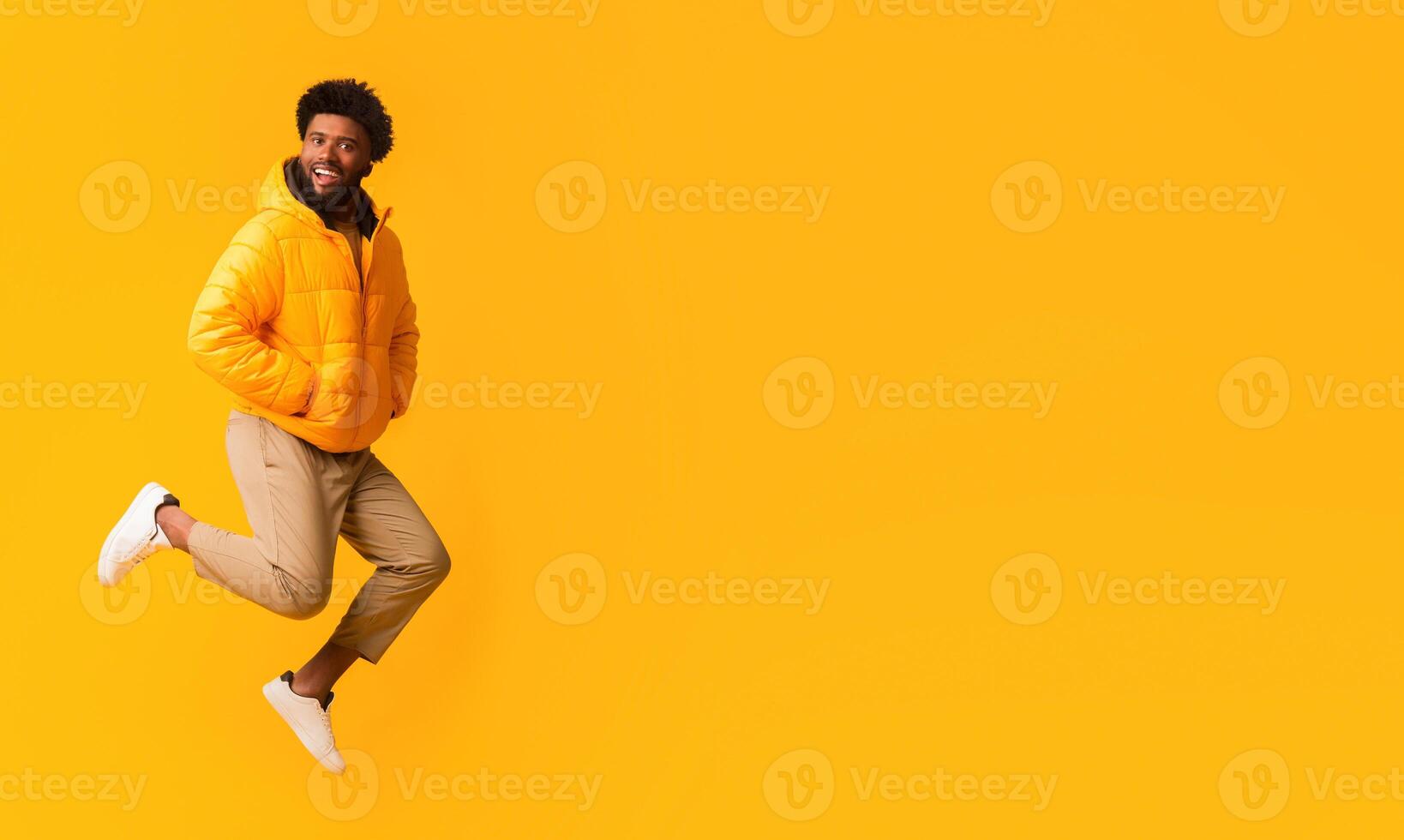 Joyful afro guy in winter jacket jumping in the air over orange background photo