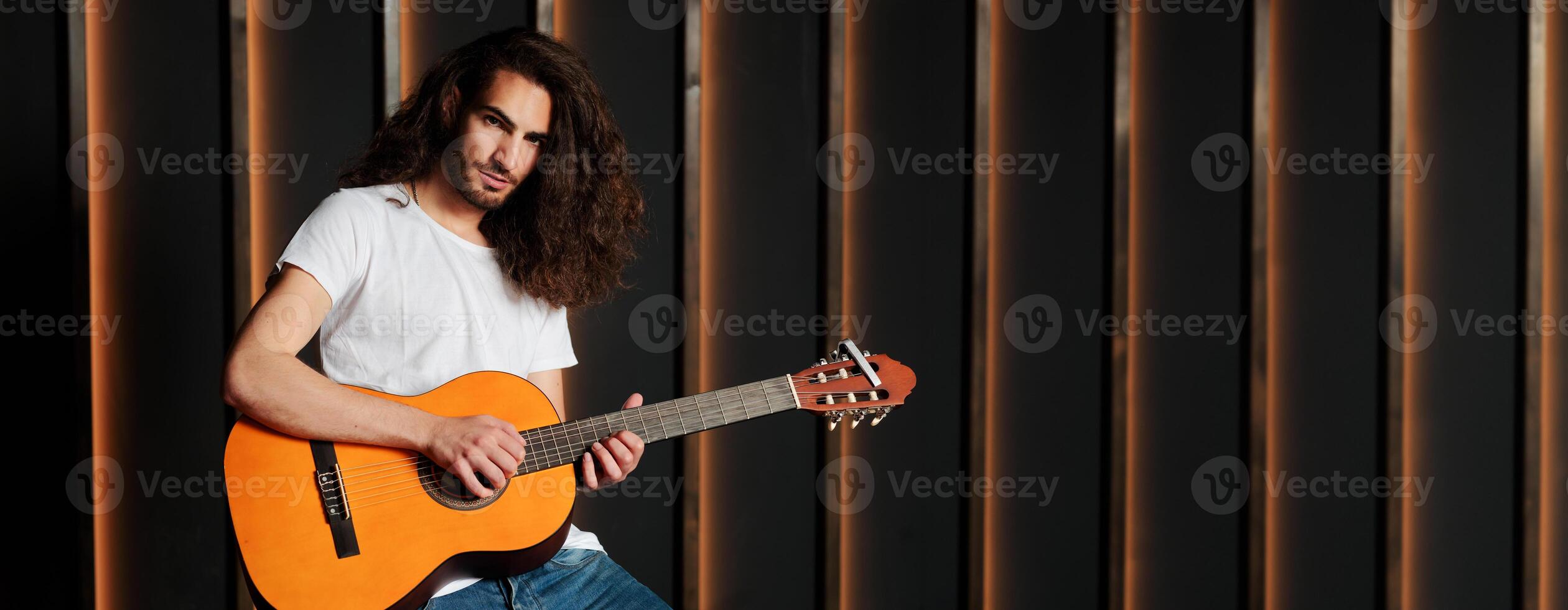 A young musician with long hair passionately strums an acoustic guitar in a modern studio setting. The warm light highlights his focused expression as he engages with his music, copy space photo