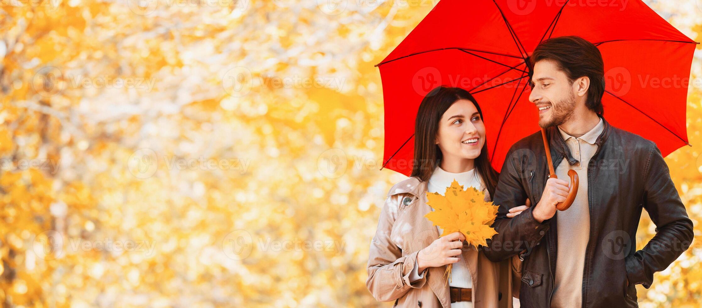 Happy young lovers with red umbrella and bunch of golden marple leaves enjoying date at autumn forest, free space photo