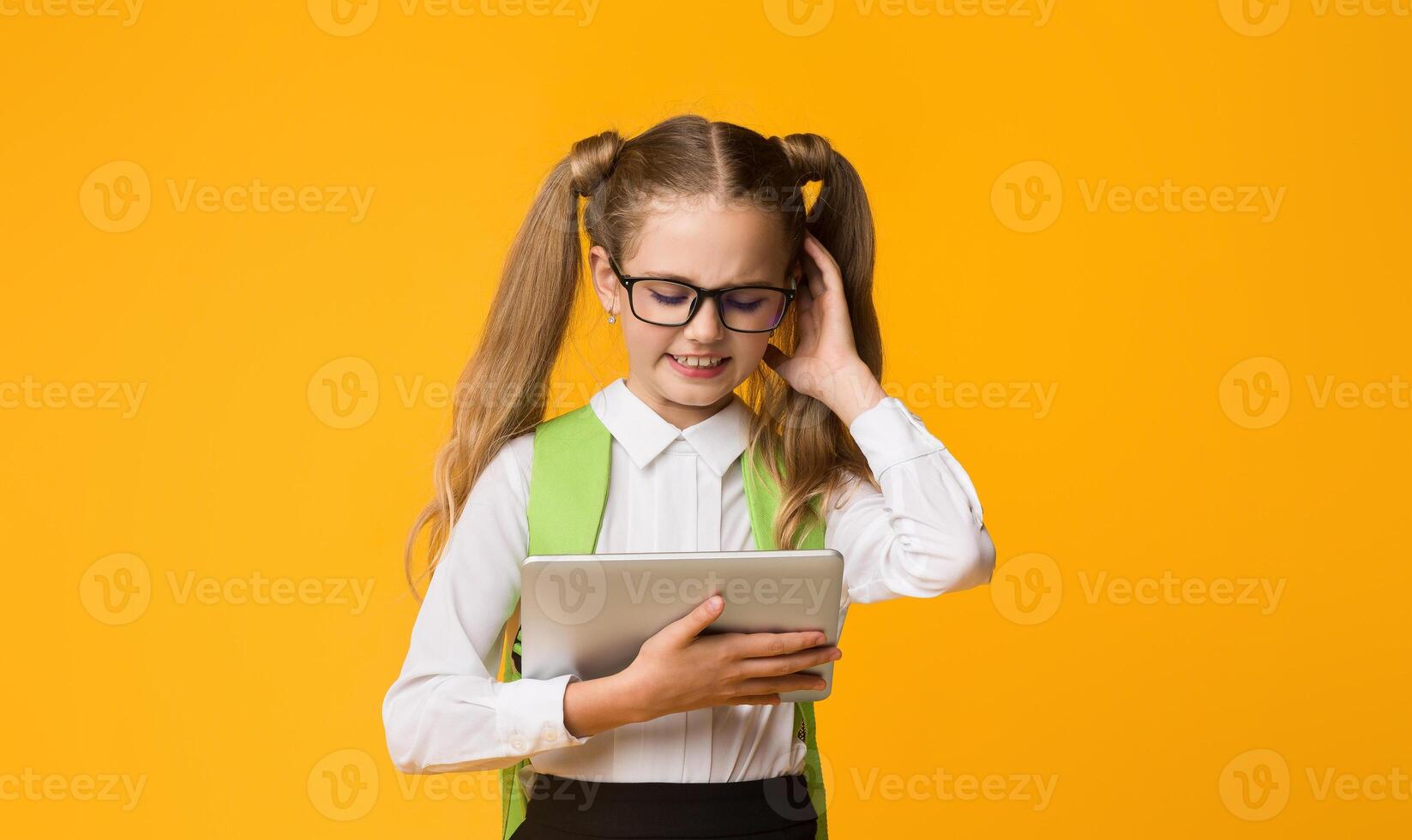 Embarassed Primary School Student Girl Scratching Head Using Digital Tablet On Yellow Background. Studio Shot, Empty Space photo
