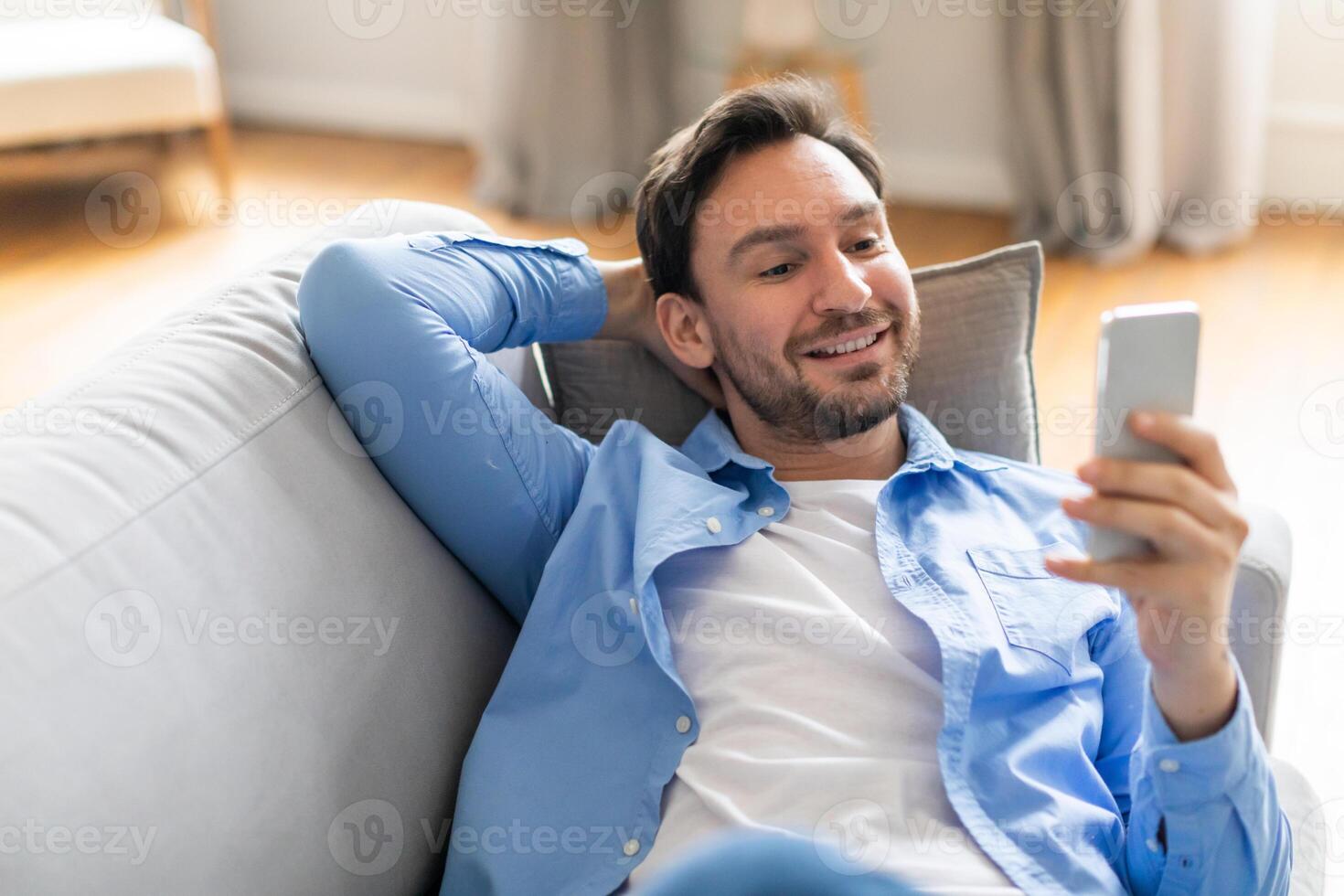 A man is seated on a couch, engrossed in his phone screen. He appears focused as he scrolls and interacts with the device. photo