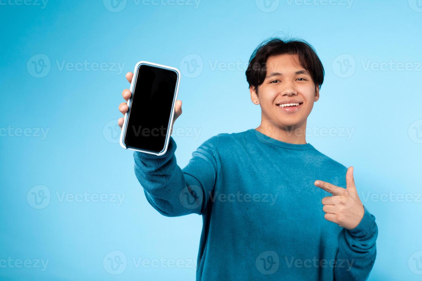 Asian smiling young guy dressed in a blue sweater points to his smartphone while standing against a solid light blue background. photo