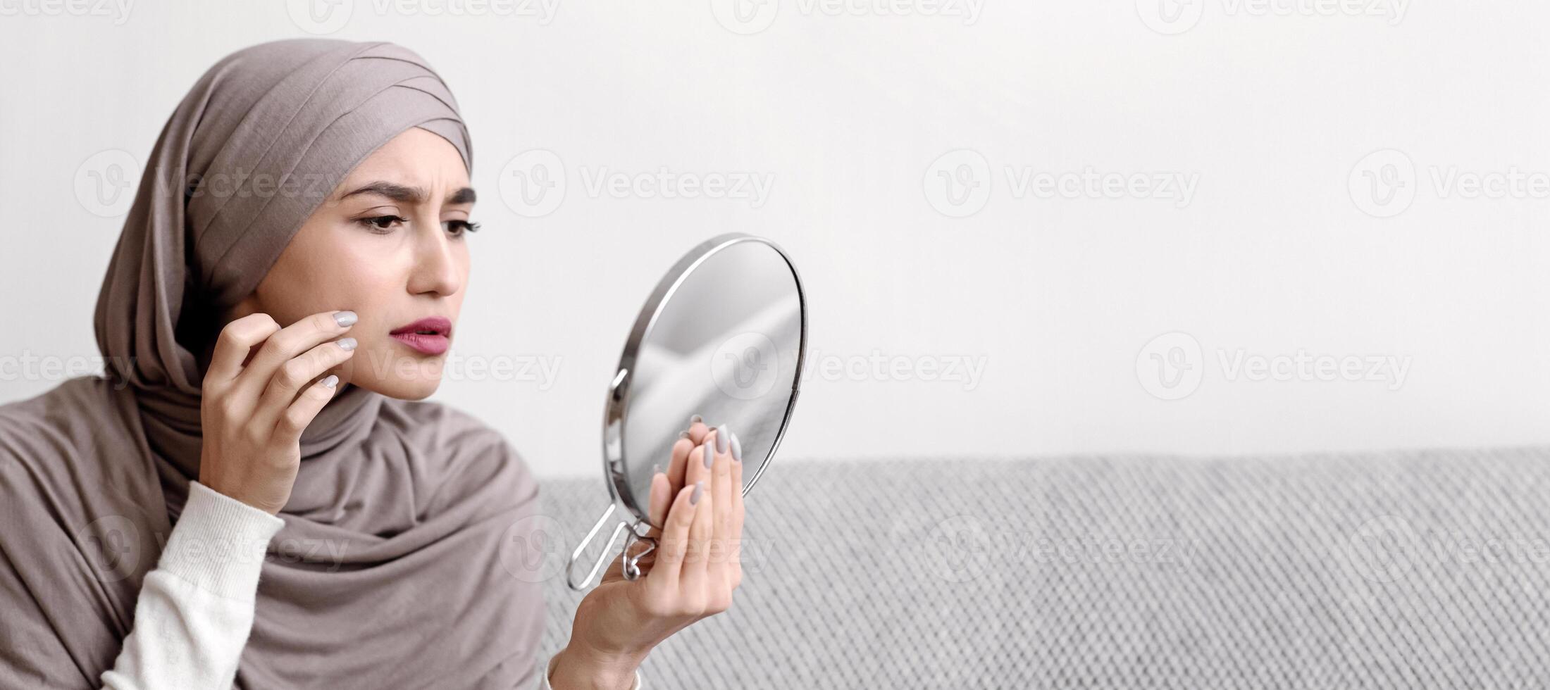 Problem Skin Concept. Muslim Girl In Hijab Looking In Round Mirror And Touching Face, Concerned About Wrinkle Or Pimple, Closeup photo