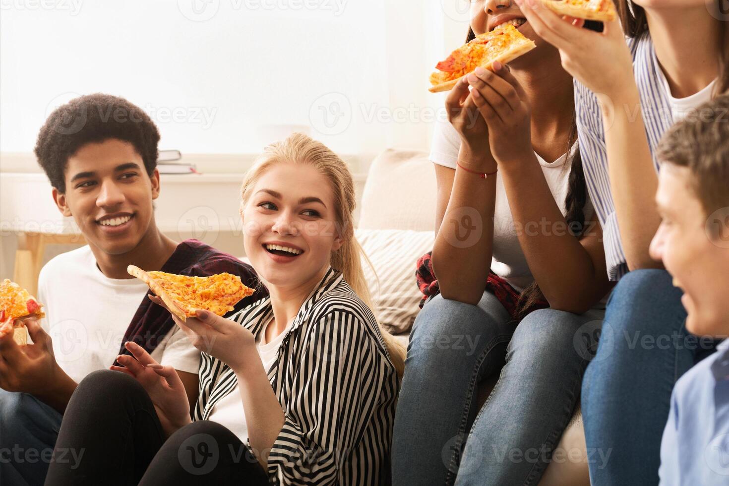 A cheerful group of young friends sits comfortably on the floor and sofa in a well-lit living room, sharing slices of pizza and engaging in lively conversation photo