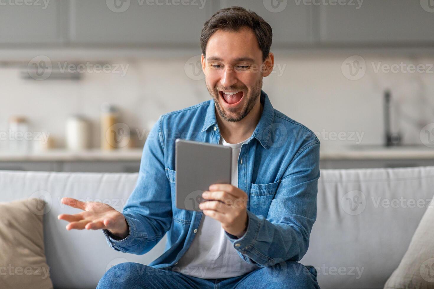 A man sits on a couch in a living room, looking excitedly at a tablet in his hands. He smiles and looks surprised as he reads the screen. photo
