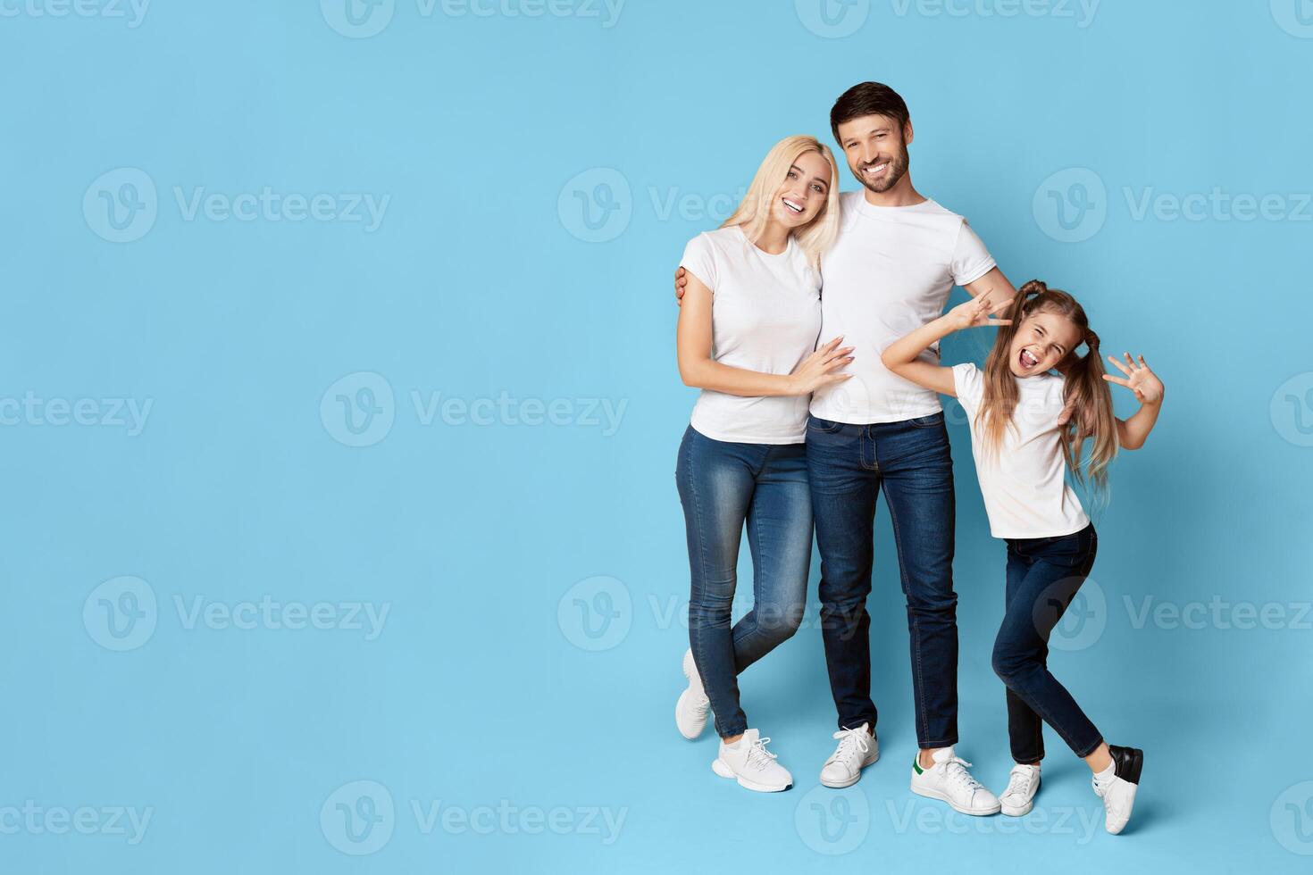 A family of three stands in front of a light blue background. The mother and father are standing close together, while the daughter stands slightly to the right, copy space photo