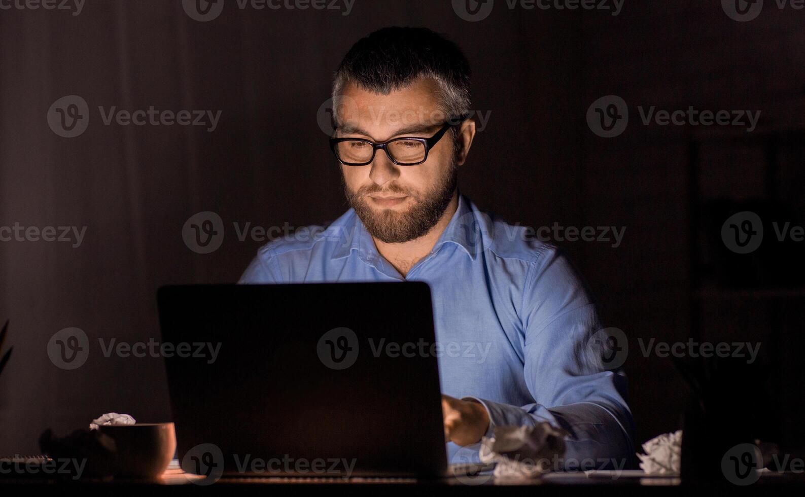 A bearded man with glasses is focused on his laptop, working late at night in a dimly lit home office, surrounded by papers. photo
